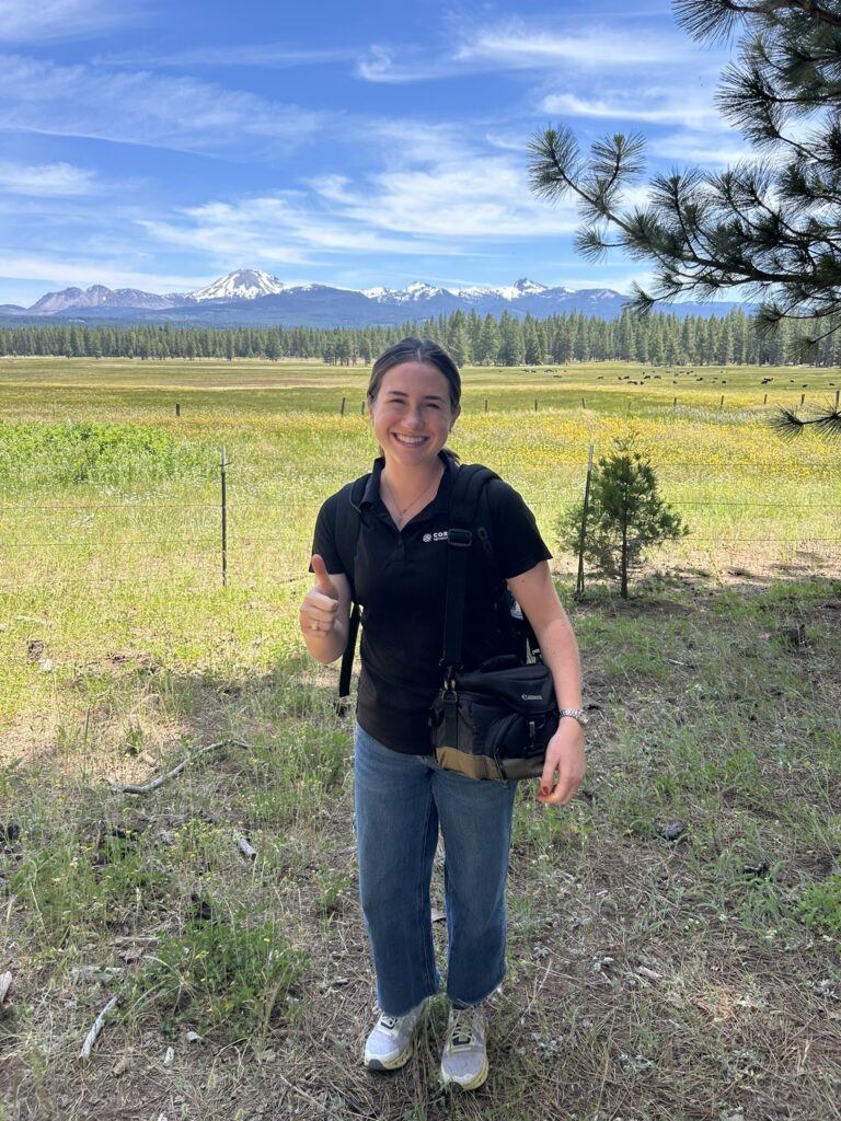 A woman stands giving a thumbs up in front of a field with the mountains in the background. 