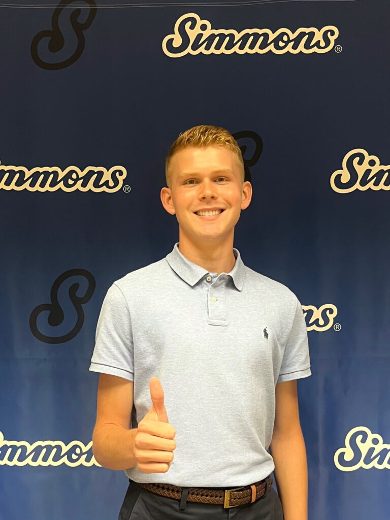 A man wearing a light blue polo shirt stands in front of a dark blue Simmons backdrop while holding his thumb up. 