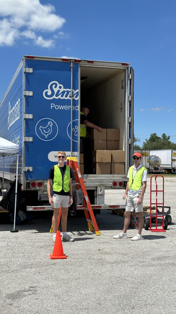 Two men wearing safety vests stand at the back of a Simmons semi-truck trailer with a cone, ladder, boxes and dolly. 