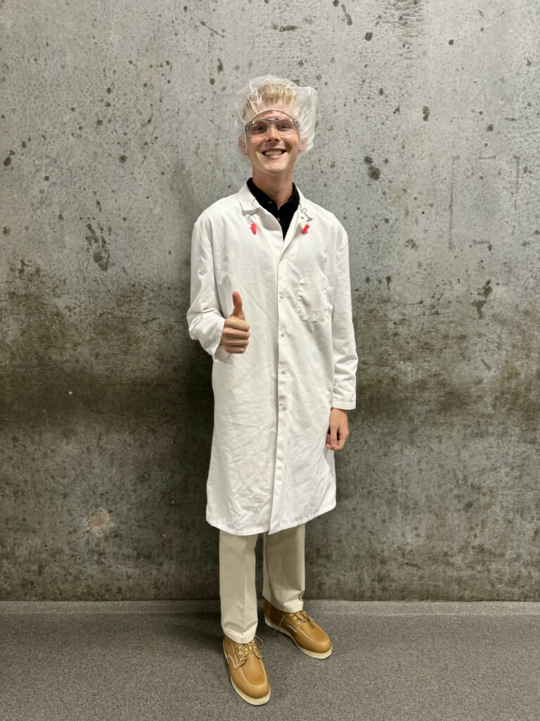 A man wearing a long white lab coat and a plastic hair net stands with his thumb up in front of a concrete wall. 