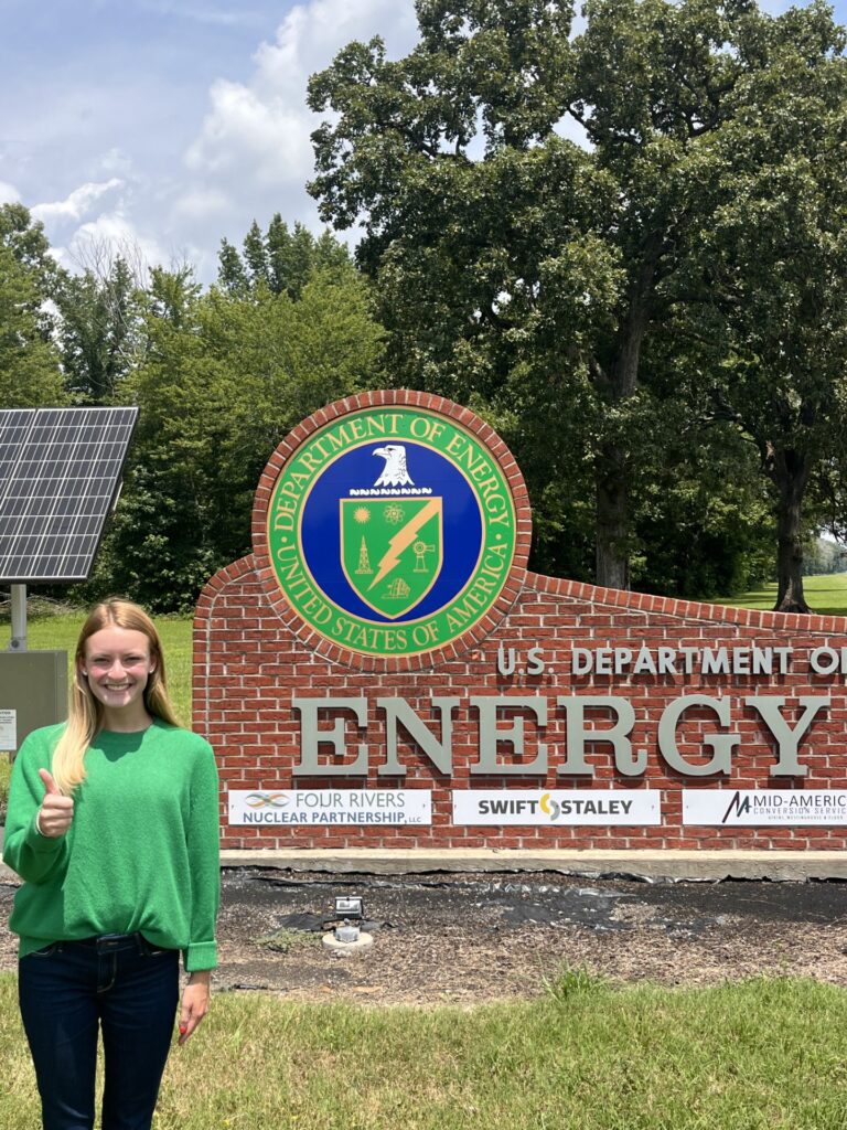 A woman in a green sweatshirt and dark pants stands with her thumb up in front of a sign for the U.S. Department of Energy. 