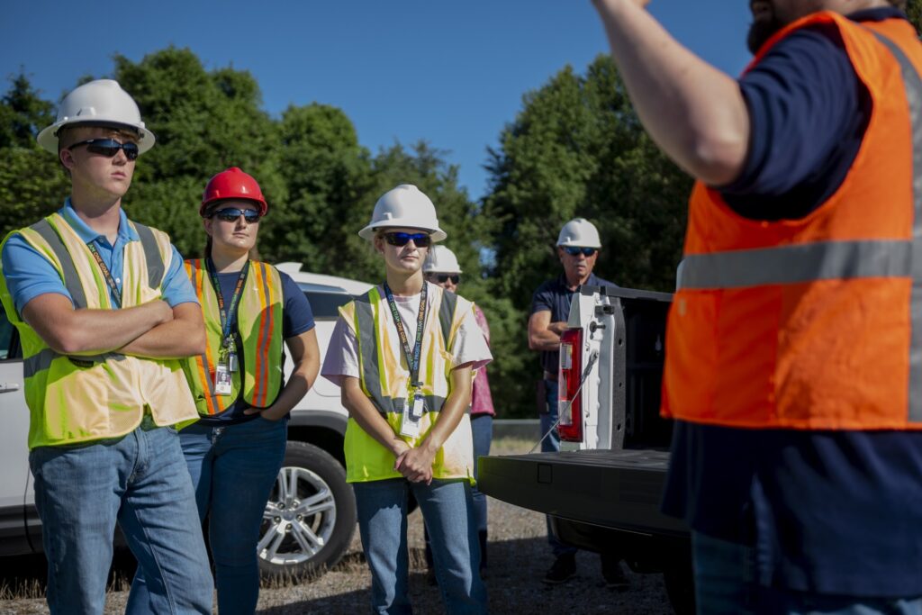 A woman in a white hard hat, sunglasses and a safety vest stands with her hands folded in front of her. 