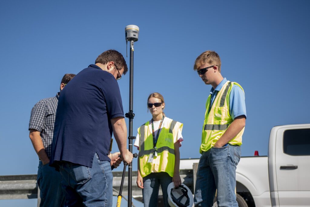 A man adjusts a black pole while a man and a woman wearing sunglasses and safety vests watch. 