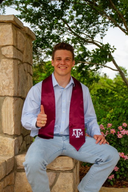 Carter Monrad '24, the first financial planning degree graduate at Texas A&M University, sits with his thumb up while wearing a light blue button up shirt, a maroon Texas A&M graduation stole and blue jeans.