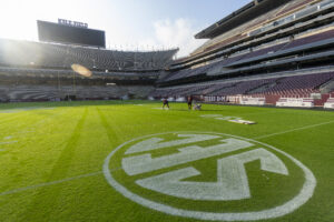 Kyle Field at Texas A&M University, with a clear morning sky, as two workers apply fresh white paint to the SEC logo on the green turf. The stadium's large, empty stands and scoreboard loom in the background, emphasizing the preparation for an upcoming football game.