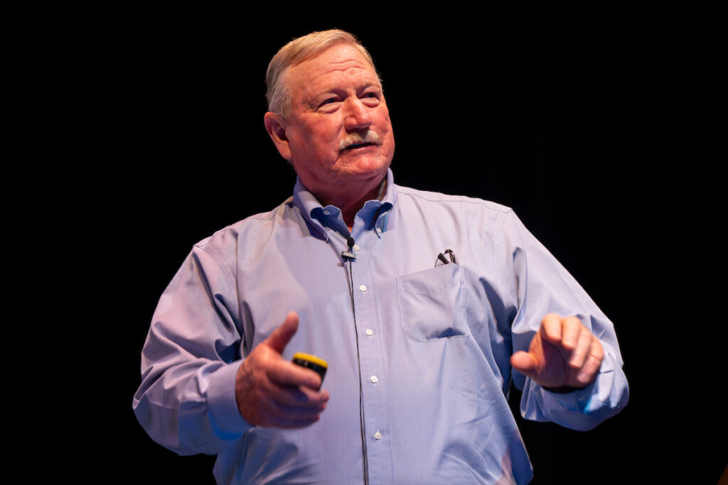 a man, Ron Gill, stands with his hands out in front against a black background and speaks on replacing animals in the beef herd