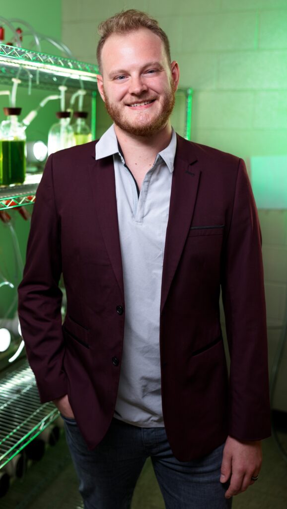 Devon Boland, Ph.D., stands in a laboratory with green-lit equipment in the background.