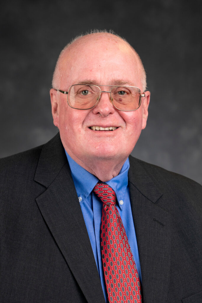 Head and shoulder photo of Bruce McCarl. Ph.D. in dark blue jacket, blue shirt and red tie. 