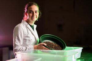 A woman in a lab coat stands confidently, holding a bowl filled with black solider flies, showcasing her research.