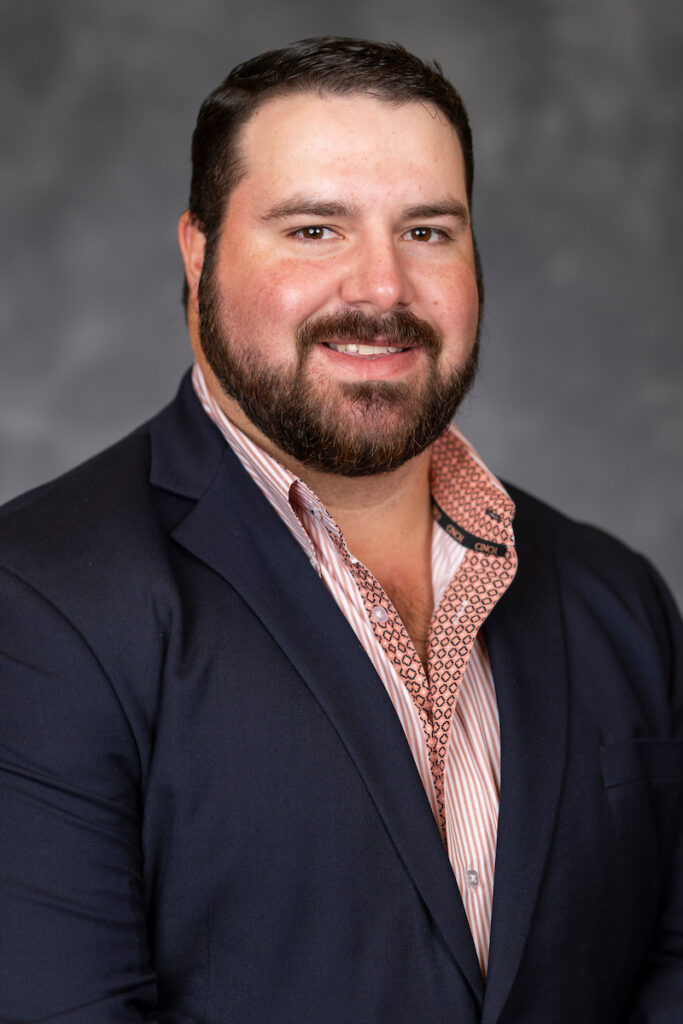 A smiling man with a beard and mustache dressed in dark jacket and pink shirt