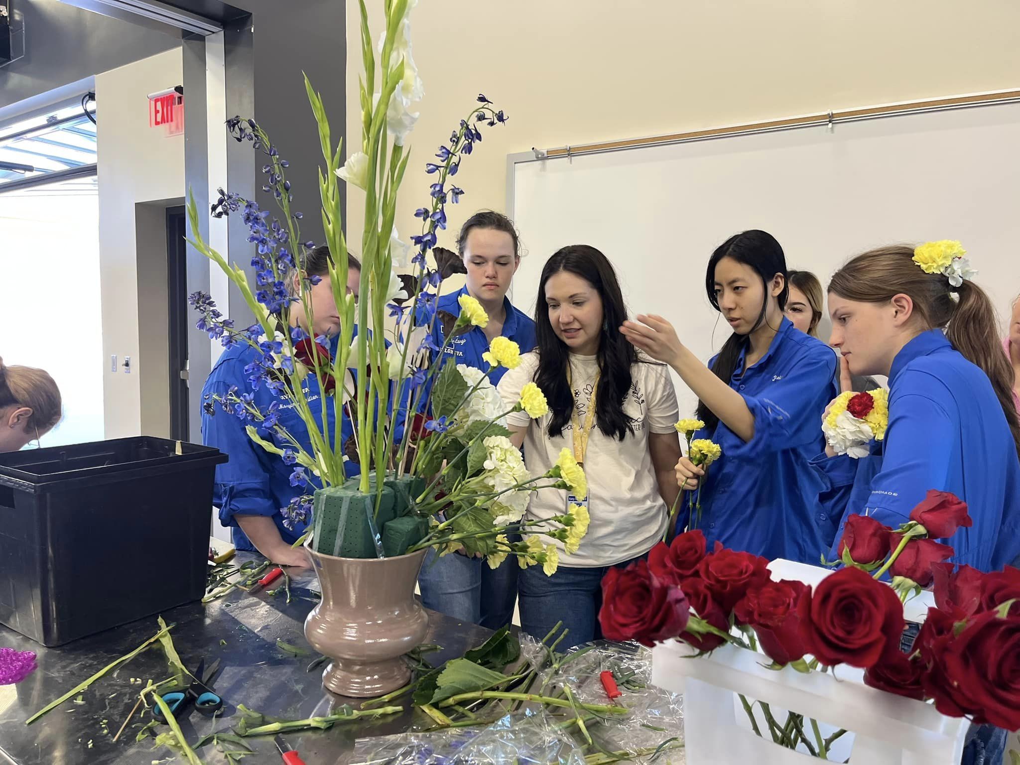 A young teacher with long dark hair instructs her students on floral design. A large floral display in in front of thenm.