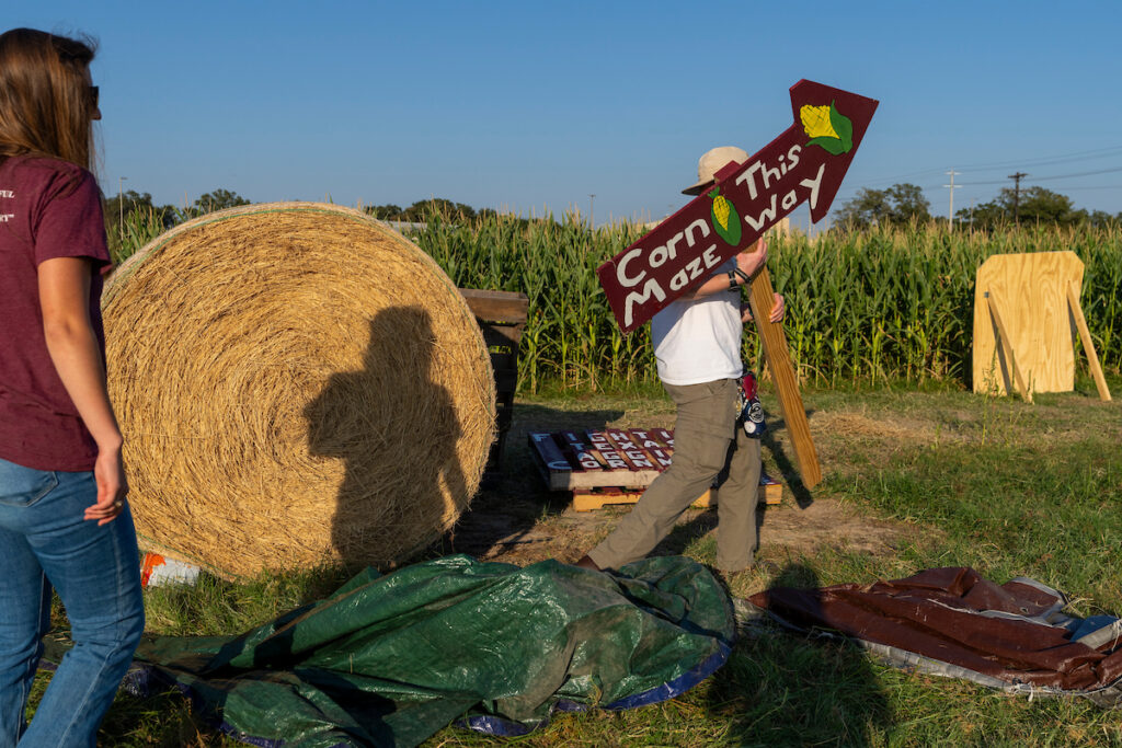 The students carry a large sign that reads “Corn Maze This Way” and the corn harvest with a large bale of hay can be seen in the background