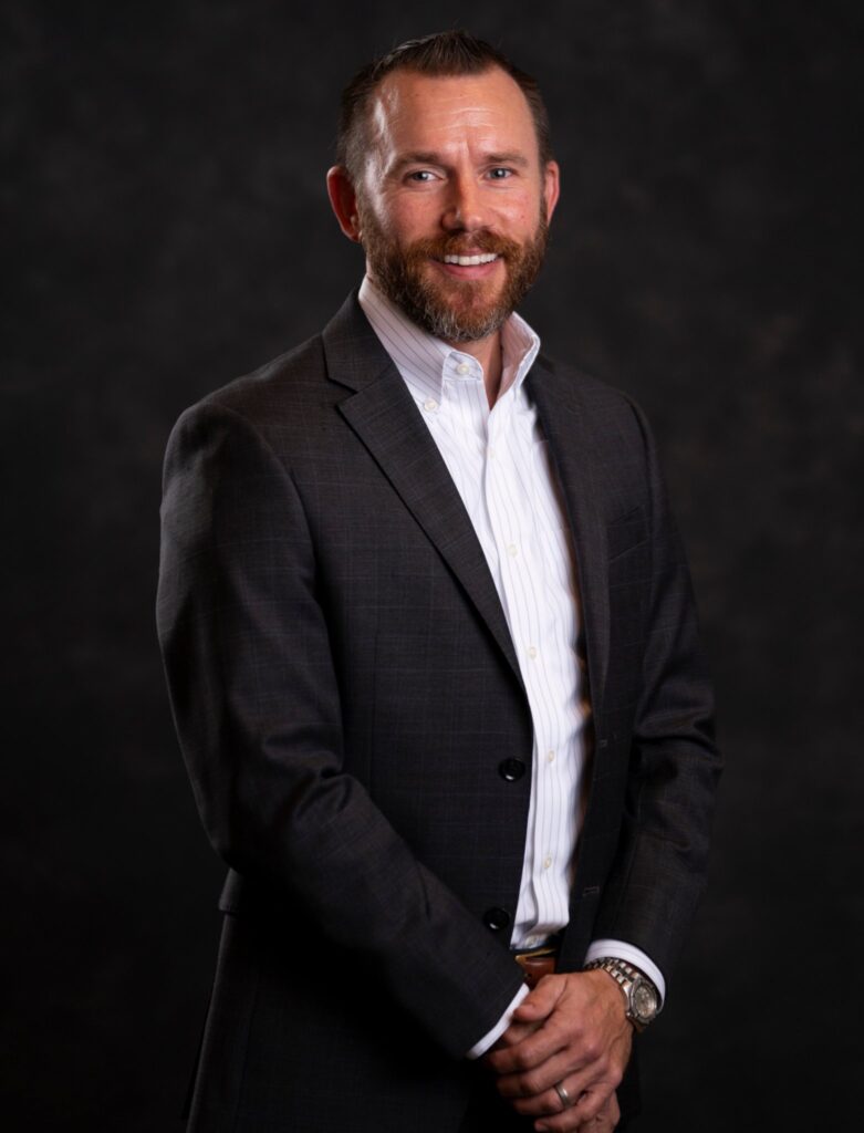 Wes Moorehead, a man with facial hair stands in a suit jacket with white shirt, no tie